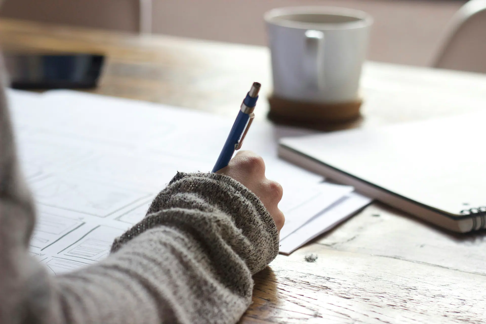 Writing notes by hand at a wooden table beside a mug